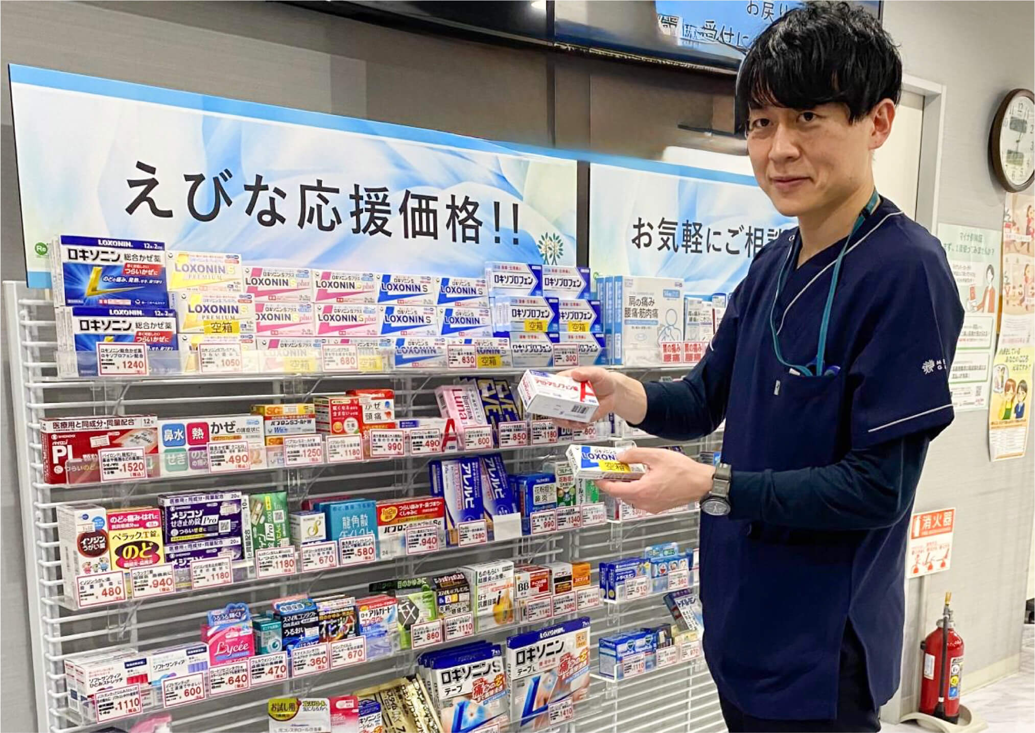 店頭の陳列棚の前でOTC医薬品を手に持ち、笑顔でご案内する男性薬剤師